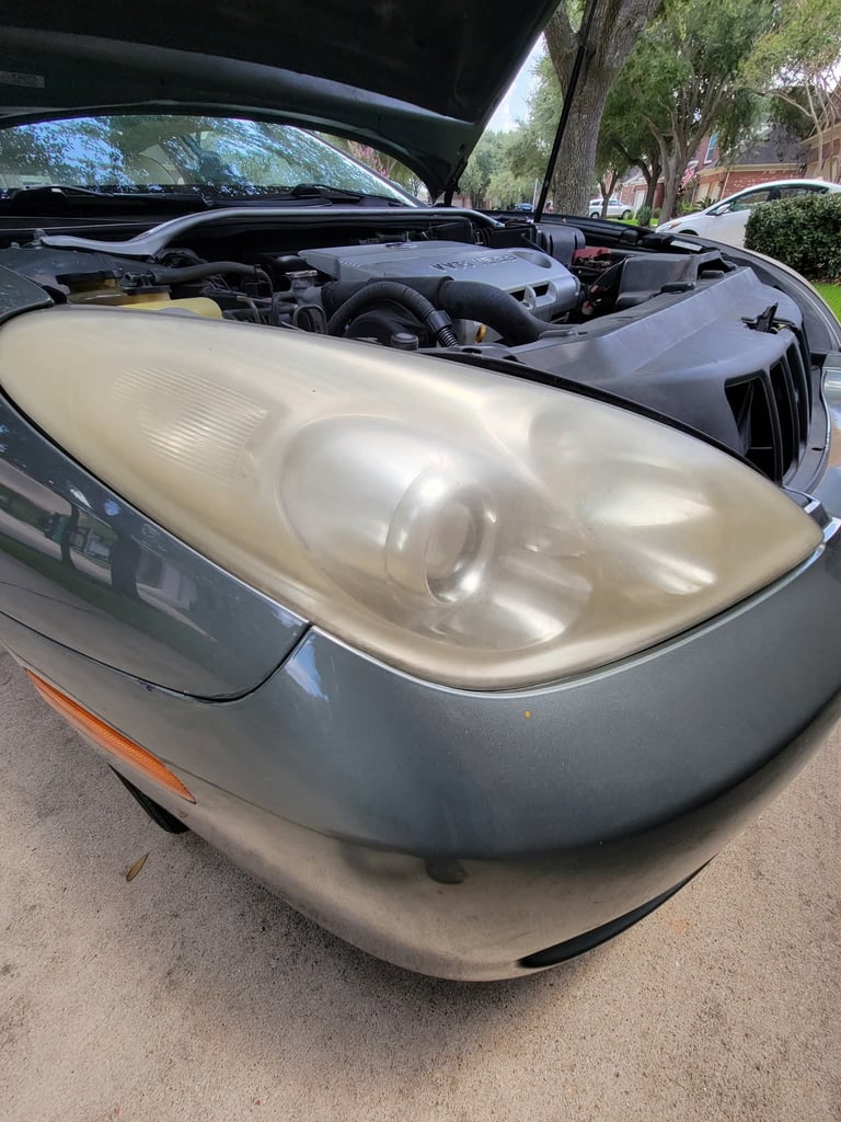 Car hood open revealing hazed headlight and engine compartment with mounted components